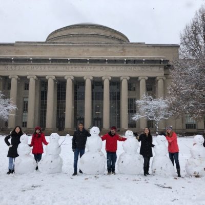 The picture shows seven people standing on the snow covered ground with arms around six snowmen. The Great Dome of MIT's Building 10 is in the background.