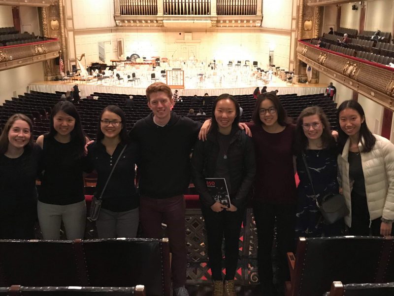 The picture shows eight people smiling and looking towards the camera with arms around each other. One is holding a program labeled BSO and the stage of Boston Symphony Hall is behind them. Teresa is third from the left with dark hair and glasses, smiling.