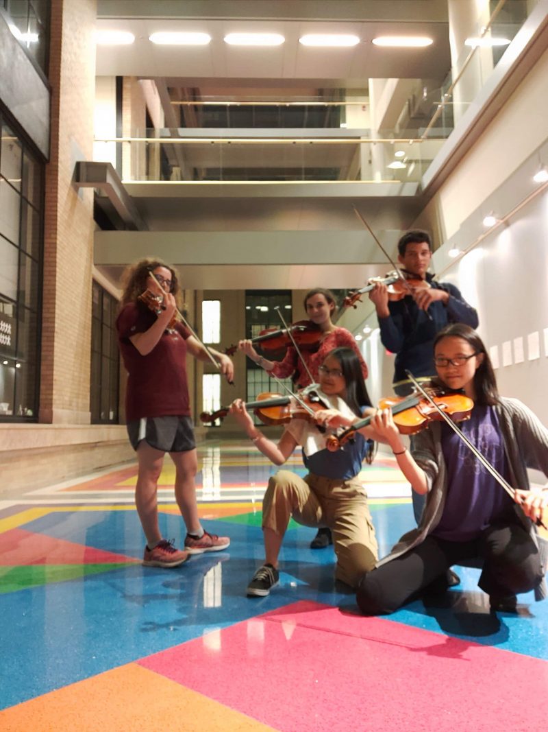 The picture shows five people standing in the lobby of an academic building, each holding a viola with their bow poised and ready to play. Teresa is kneeling in the front center of the photo, smiling with long dark hair and glasses.